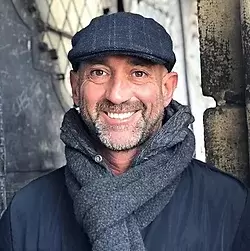 Smiling man in a flat cap and scarf outdoors.