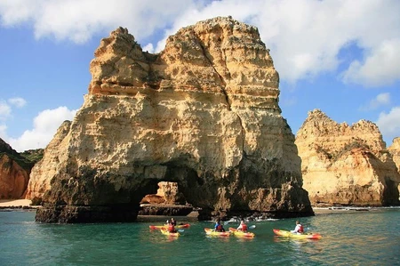 Kayakers paddling near large coastal cliffs with caves under a blue sky.