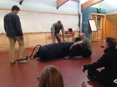 Three men handling a large black object indoors with onlookers.