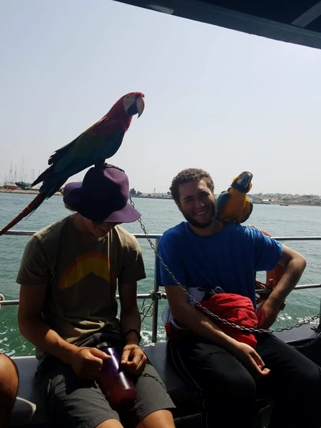 Two men sitting on a boat, one wearing a parrot on his hat.