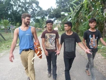 Four young men walking outdoors along a rural road, surrounded by greenery.