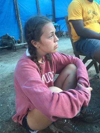 A young woman in a pink hoodie sitting outdoors, looking to the side thoughtfully.