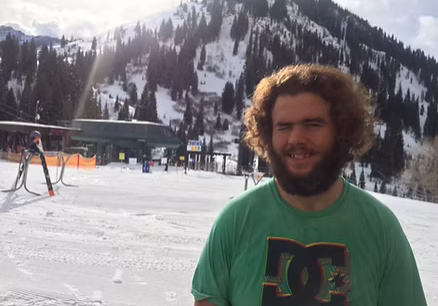 A man with curly hair and a beard smiles in a snowy mountain setting.