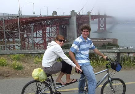 Two people riding a tandem bike near a red bridge on a foggy day.