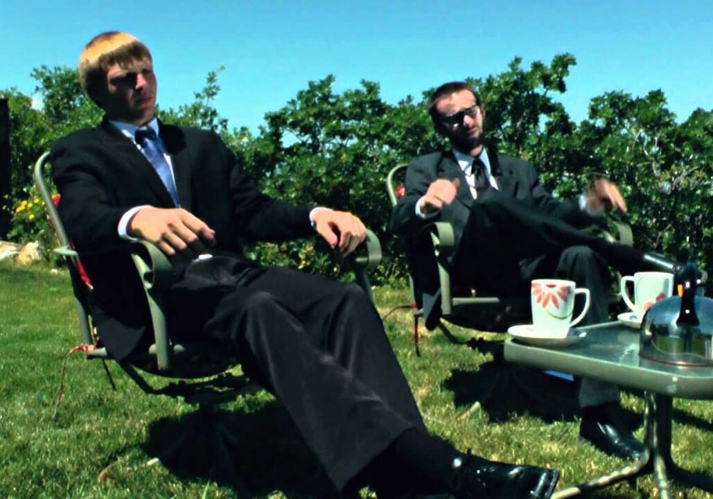Two men in suits relaxing outdoors on chairs under a sunny sky.